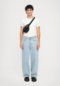 Bolsa preta a tiracolo com um bolso com fecho, usada sobre uma t-shirt branca simples e calças de ganga largas em azul claro. Calçado preto.