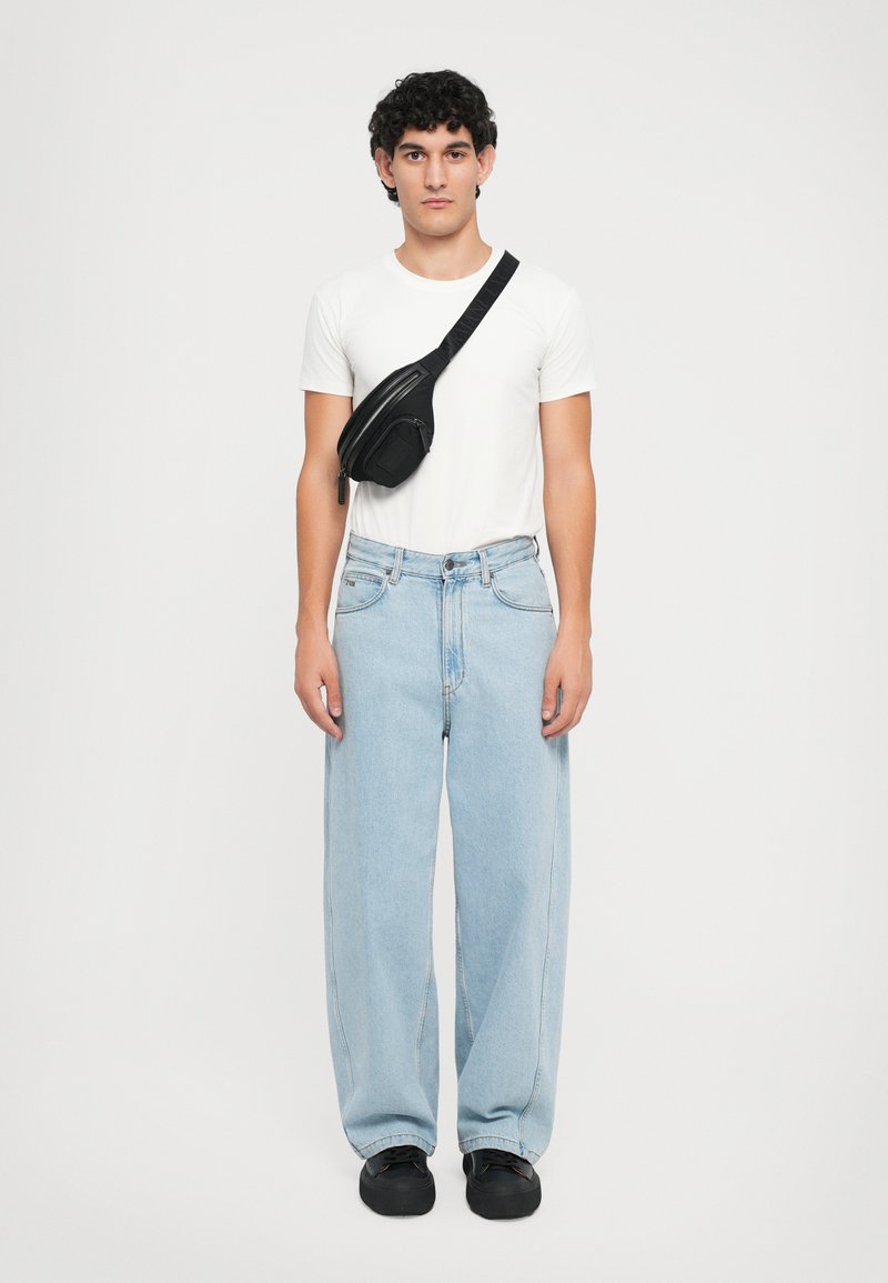 Bolsa preta a tiracolo com um bolso com fecho, usada sobre uma t-shirt branca simples e calças de ganga largas em azul claro. Calçado preto.