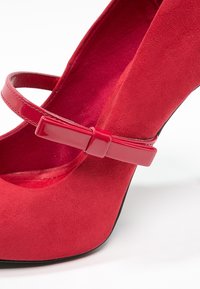 Red suede high-heeled shoe with a wide strap and a glossy bow accent. Interior is also red, and the sole is black.