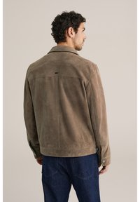 Brūns zamšādas žakete ar apkakli, redzamu šuvi un spiedpogas akcentiem. Tai ir taisna piegriezuma forma, un to valkā virs tumšām džinsām.