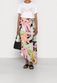 Witte t-shirt met bloemenprint, gecombineerd met een meerkleurige, asymmetrische rok met grote bladerenpatronen. Zwarte handtas en muiltjes.