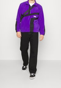 Lilla fleecejakke med sorte detaljer og lynlås. Et par sorte bukser og sorte sneakers med hvide snørebånd fuldender outfittet.
