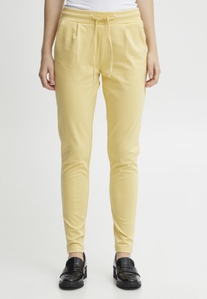 Pantalons de jogging élastiques jaunes avec taille à cordon, poches latérales et design fuselé, associés à des mocassins noirs brillants.
