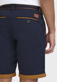 Mørkeblå shorts med brune detaljer, oppbrettede ermer og baklommer med knapp; laget av et lett stoff.