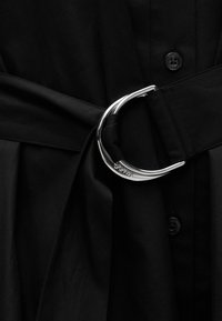 Μαύρη υφασμάτινη ζώνη με ασημένια μεταλλική αγκράφα σε σχήμα D στερεωμένη πάνω σε μαύρο ρούχο με κουμπιά.