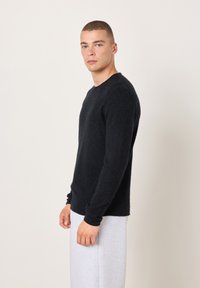 Schwarzer Strickpullover mit Rundhalsausschnitt, weicher Textur, langen Ärmeln. Das Modell trägt ihn mit hellgrauen Jogginghosen und steht vor einem neutralen Hintergrund.