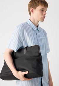 Geantă din nylon neagră, cu textură netedă, formă rectangulară, și două mânere superioare, ținută de o persoană îmbrăcată într-o cămașă albă cu dungi bleu.