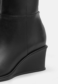 Černé kotníkové boty na klínu vyrobené z texturované kůže. Mají zakřivený klínový podpatek a hladký steh podél švu.