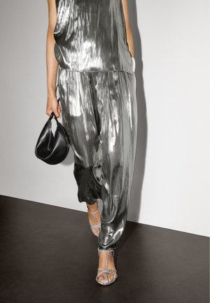 Top e pantaloni metallici argento con una texture lucida; borsa nera; sandali argentati con cinturini. Design semplice e fluido con pieghe.