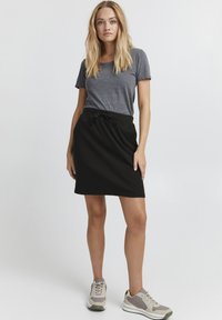T-shirt gris à manches courtes, associé à une jupe noire jusqu'au genou avec un lien à la taille. Le mannequin porte des baskets gris clair avec des accents verts.