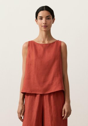 Vrouw draagt een mouwloos loszittend linnen topje in roestkleur en bijpassende broek met wijde pijpen, staand tegen een effen lichte achtergrond.