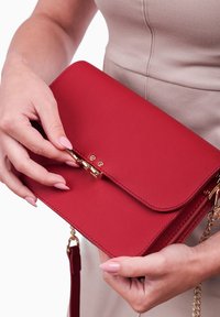 Červená kabelka z falošnej kože s klopou, zlatými prvky a odnímateľným ramenním popruhem. Disponuje hladkým povrchom a strukturovaným designem.