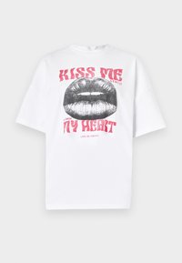 T-shirt bianco oversized con una grafica di labbra nere e il testo rosso "BACIAMI GENTILMENTE" e "IL MIO CUORE" sottostante. Design semplice e casual.