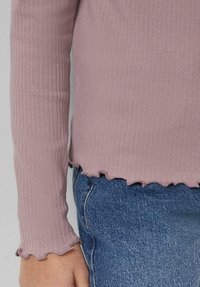 Prążkowany top z długim rękawem w bladoróżowym kolorze, z falbankowymi mankietami i dołem, zestawiony z niebieskim dżinsem. Materiał wydaje się miękki i lekko elastyczny.