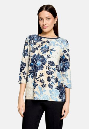 Femme aux longs cheveux foncés portant un haut crème avec un motif floral bleu et des manches trois-quarts, associée à un pantalon noir, debout sur fond blanc.