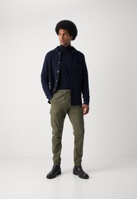 Marineblaue Strickjacke mit braunen Knöpfen, getragen über einem dunklen Rollkragenshirt, kombiniert mit olivgrünen Cargo-Hosen und schwarzen Stiefeletten.