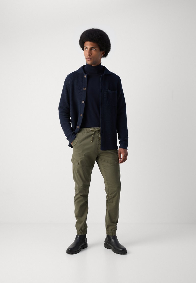 Cardigan en tricot marine avec des boutons marron, porté sur un col roulé sombre, assorti à un pantalon cargo vert olive et des bottines noires.