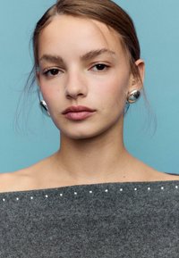 Sivo pulover z odkritimi rameni in majhnimi zaključki z zatiči na izrezu; srebrni opazni uhani. Ozadje je enobarvno pastelno modro.