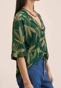 Grüne kurzärmlige Bluse mit einem goldenen Blattmuster und einem V-Ausschnitt. Getragen mit hoch taillierten blauen Denim-Jeans.