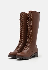 Braune High-Heel-Stiefel aus Kunstleder, mit einem hohen Schaft, goldenen Ösen und einer schnürbaren Vorderseite. Strukturierte Sohle sorgt für Grip.