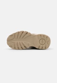 Semelle de chaussure en caoutchouc beige présentant une surface texturée avec des motifs de crampons proéminents et un logo de marque en relief, conçue pour l'adhérence et la durabilité.