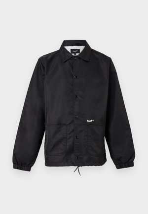 Sort skjortejakke med krave, elastiske ærmekanter, forlommer, snøre i kanten og lille hvidt "huf"-logo på venstre forside.