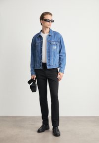 Hellblaues Jeansjackett mit Fronttaschen, graues T-Shirt, schwarze Hose und schwarze Schuhe. Hält eine Kamera und trägt dunkle Sonnenbrillen.
