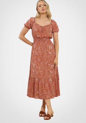 Kobieta w czerwonej sukience midi w kwiaty z bufiastymi rękawami, stojąca z jedną ręką na biodrze, w brązowych wsuwanych sandałach na jednolitym tle.