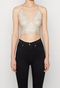 Metalická béžová crop top se hlubokým výstřihem do V, s nařasenou strukturou a zavazováním za krkem, doplněná černými džínami s vysokým pasem.