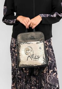 Mochila negra y dorada hecha de material texturizado con un diseño gráfico que presenta una figura y texto. Tiene dos asas en la parte superior y cierre de cremallera.