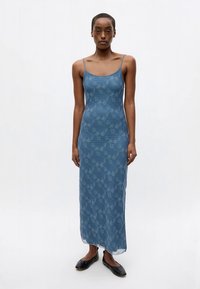 Mulher vestindo um vestido maxi azul sem mangas com um padrão floral discreto e sapatilhas pretas, de pé contra um fundo branco liso.