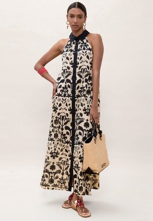 Mujer con vestido largo sin mangas de flores negro y beige con cuello negro, sosteniendo un bolso tote color canela, lleva una pulsera roja, pendientes y sandalias con flores rojas.