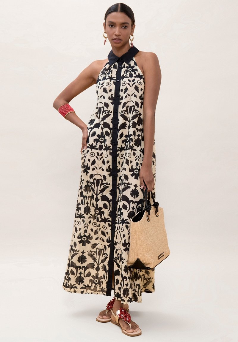 Mujer con vestido largo sin mangas de flores negro y beige con cuello negro, sosteniendo un bolso tote color canela, lleva una pulsera roja, pendientes y sandalias con flores rojas.