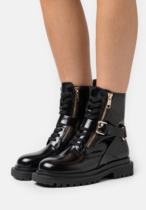 Stivaletti alla caviglia in pelle verniciata nera con doppia cerniera, allacciatura frontale, dettaglio con fibbia, suola chunky e minuteria dorata a contrasto.