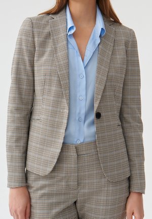 Woman wearing a tailored brown and white checkered blazer and matching pants over a light blue button-up shirt.