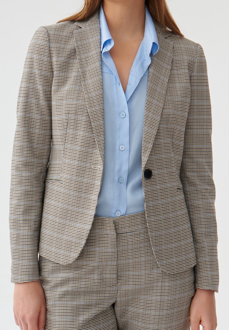 Femme portant un blazer marron et blanc à carreaux sur mesure et un pantalon assorti, par-dessus une chemise bleu clair boutonnée.