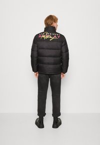 Schwarze Pufferjacke mit einem gesteppten Design, auf der Rückseite mit buntem Text. Trägt sie zu dunklen Jeans und klobigen schwarzen Stiefeln.