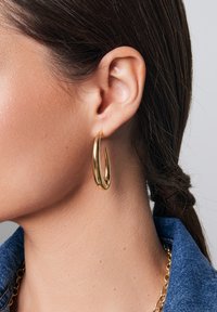 Złote kolczyki w formie obręczy o smukłym, wydłużonym kształcie i wypolerowanej powierzchni. Noszone na uchu, uzupełniające dżinsową kurtkę.