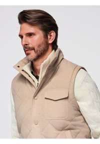 Gilet trapuntato beige con colletto alto, tasca frontale e chiusure a scatto, indossato sopra un maglione beige a trecce. Texture liscia.