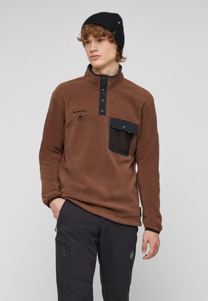 Junger Mann trägt einen braunen Pullover mit schwarzen Druckknöpfen und Tasche, eine schwarze Mütze und schwarze Hose mit seitlicher Reißverschlusstasche.