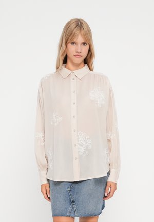 Camicia button-up leggera beige chiaro e trasparente con maniche lunghe, caratterizzata da ricami floreali bianchi sulle spalle e un collo classico. Gonna di denim.