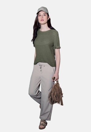 Kobieta ubrana w oliwkowozielony sweter, beżowe spodnie, beżową czapkę i sandały, trzymająca w lewej ręce brązową torebkę z frędzlami.