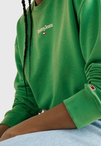 Osoba v zelené mikině Tommy Jeans s logem na hrudi a rukávu, opírá ruku o světle modrou džínovou tkaninu.