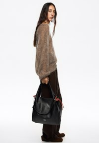 Czarna skórzana torba hobo o fakturowanym wykończeniu, z czerwonymi ozdobnymi sznurkami i miękkim, luźnym kształtem; trzymana przez osobę ubrana w miękki, kremowy sweter.