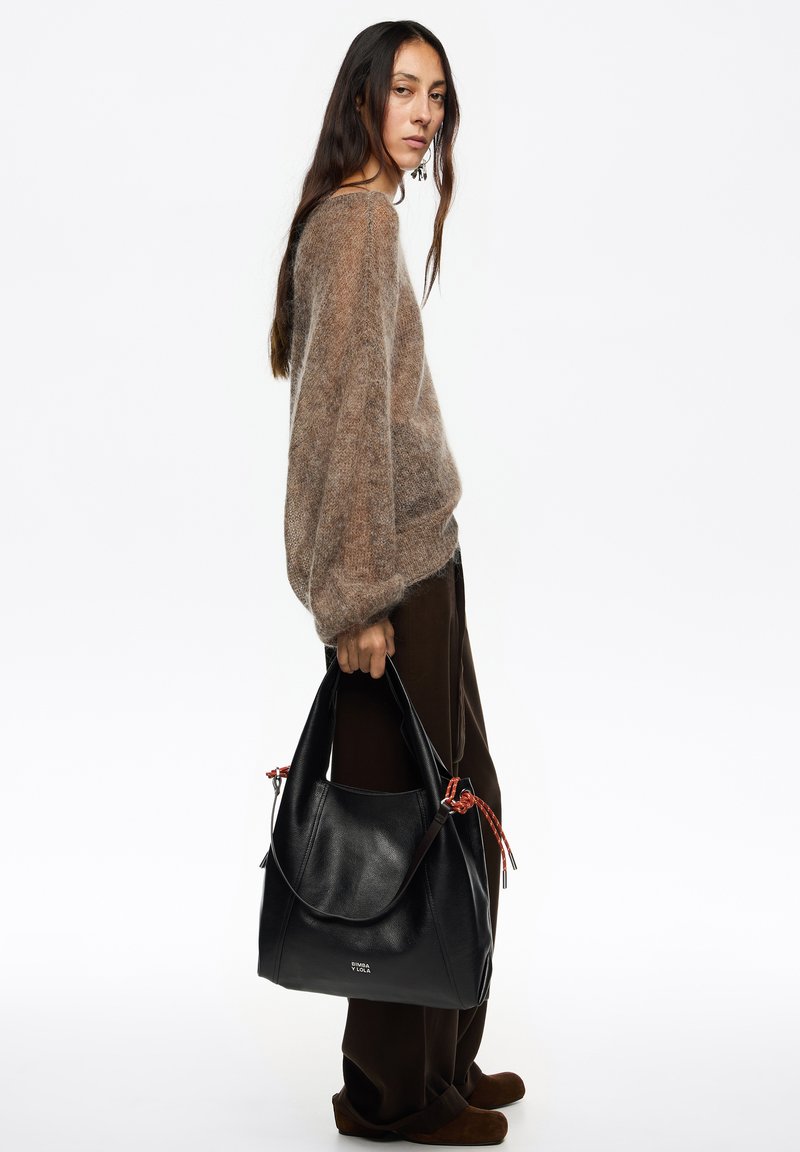 Czarna skórzana torba hobo o fakturowanym wykończeniu, z czerwonymi ozdobnymi sznurkami i miękkim, luźnym kształtem; trzymana przez osobę ubrana w miękki, kremowy sweter.