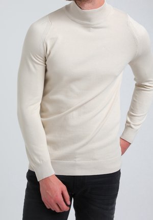 Homme portant un pull à col roulé crème ajusté avec des poignets et un ourlet côtelés, associé à un pantalon noir, sur un fond uni.