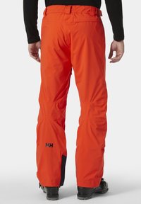 Heren oranje skibroek van waterdicht materiaal. Voorzien van een tailleband met riemlussen en een logo op de dij. Verstelbare boorden.