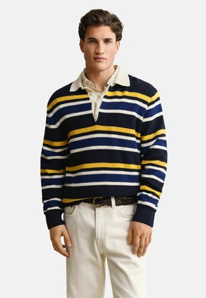 Man draagt een rugbyshirt met strepen in marineblauw, geel, wit en blauw over een beige overhemd, gecombineerd met off-white broek en een bruine gevlochten riem.