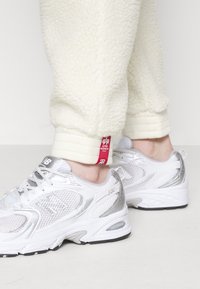 Silberne und weiße Sportschuhe mit Netzpaneelen und reflektierenden Akzenten; kombiniert mit creamfarbigen Fleece-Jogginghosen, die ein rotes Etikett am Saum haben.