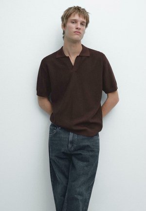 Giovane uomo con capelli biondi corti che indossa una maglietta a maniche corte marrone scuro e jeans neri sbiaditi, in piedi contro un muro bianco.